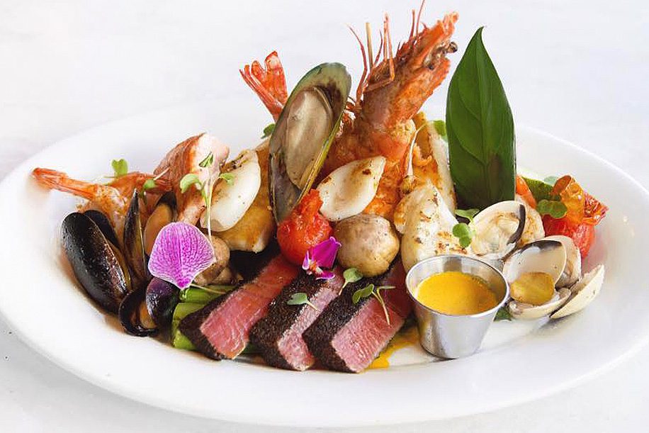 A plate of seafood and steak with a small bowl of sauce.