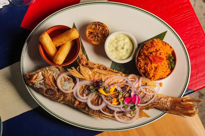A plated dish of whole fish with onions and flowers on top, served with rice, fried rolls, and a small bowl of sauce.