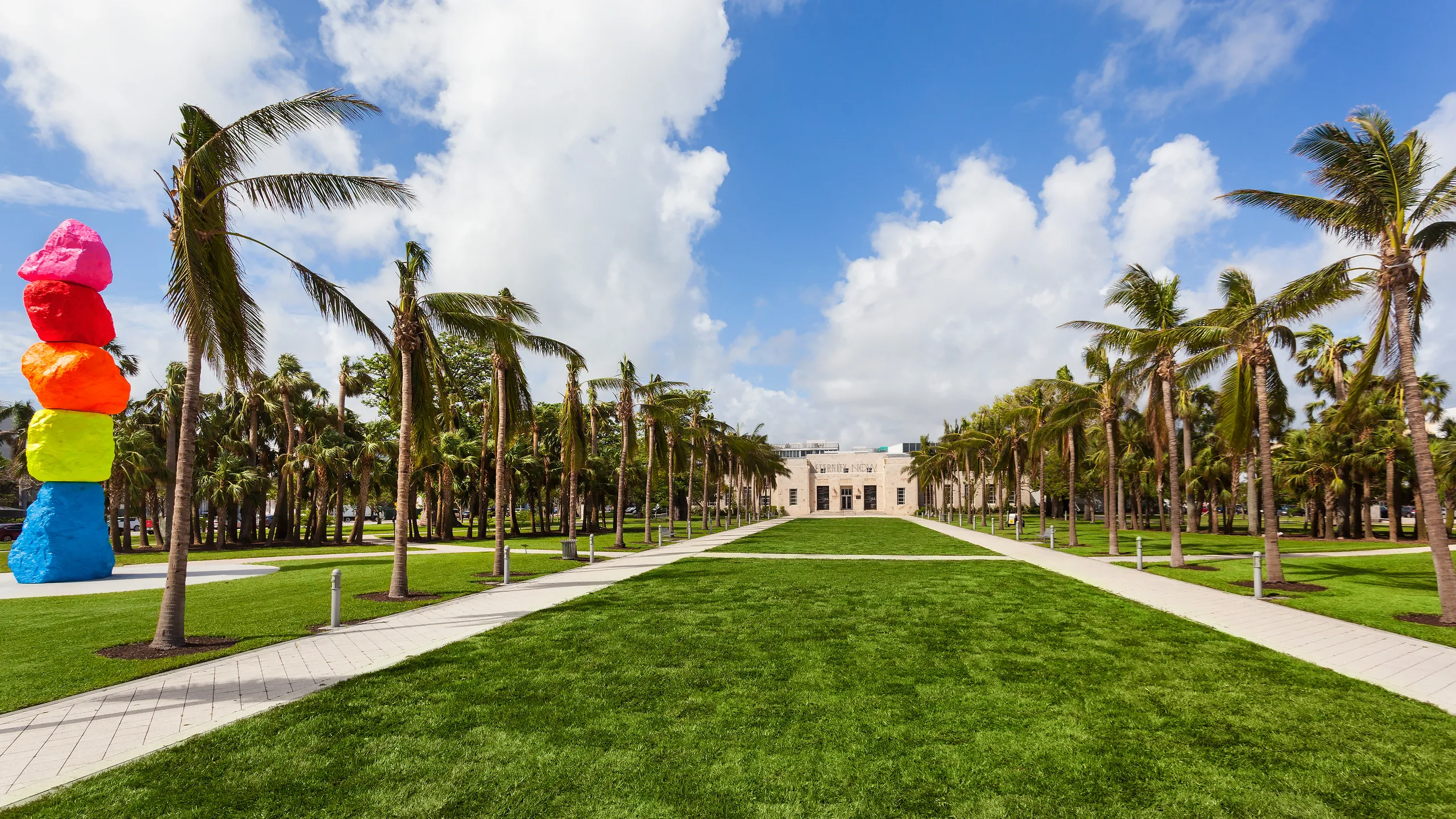 A wide shot of a building with a colorful sculpture in front of it, surrounded by palm trees and grass.