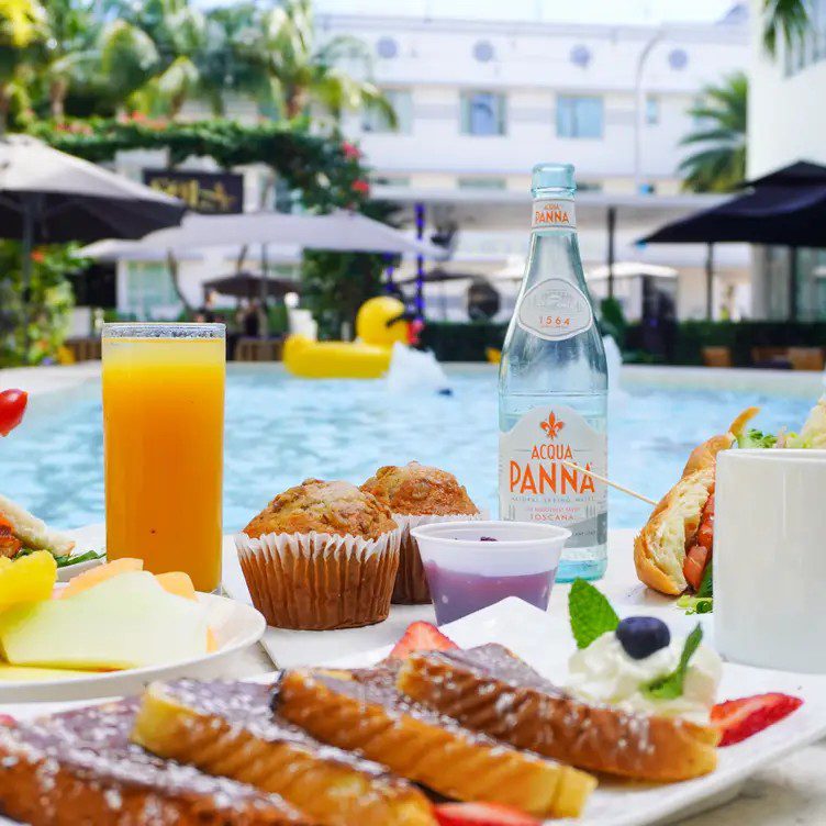 A spread of breakfast foods and drinks on a table in front of a pool with a large white building in the background.