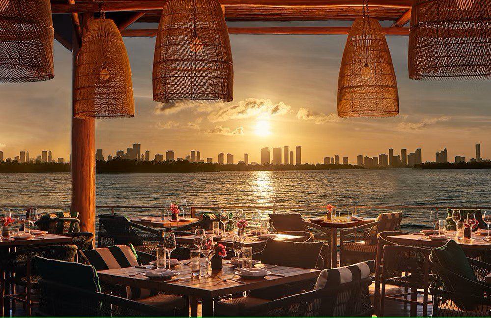 A restaurant with outdoor seating and a view of the water and city skyline at sunset.