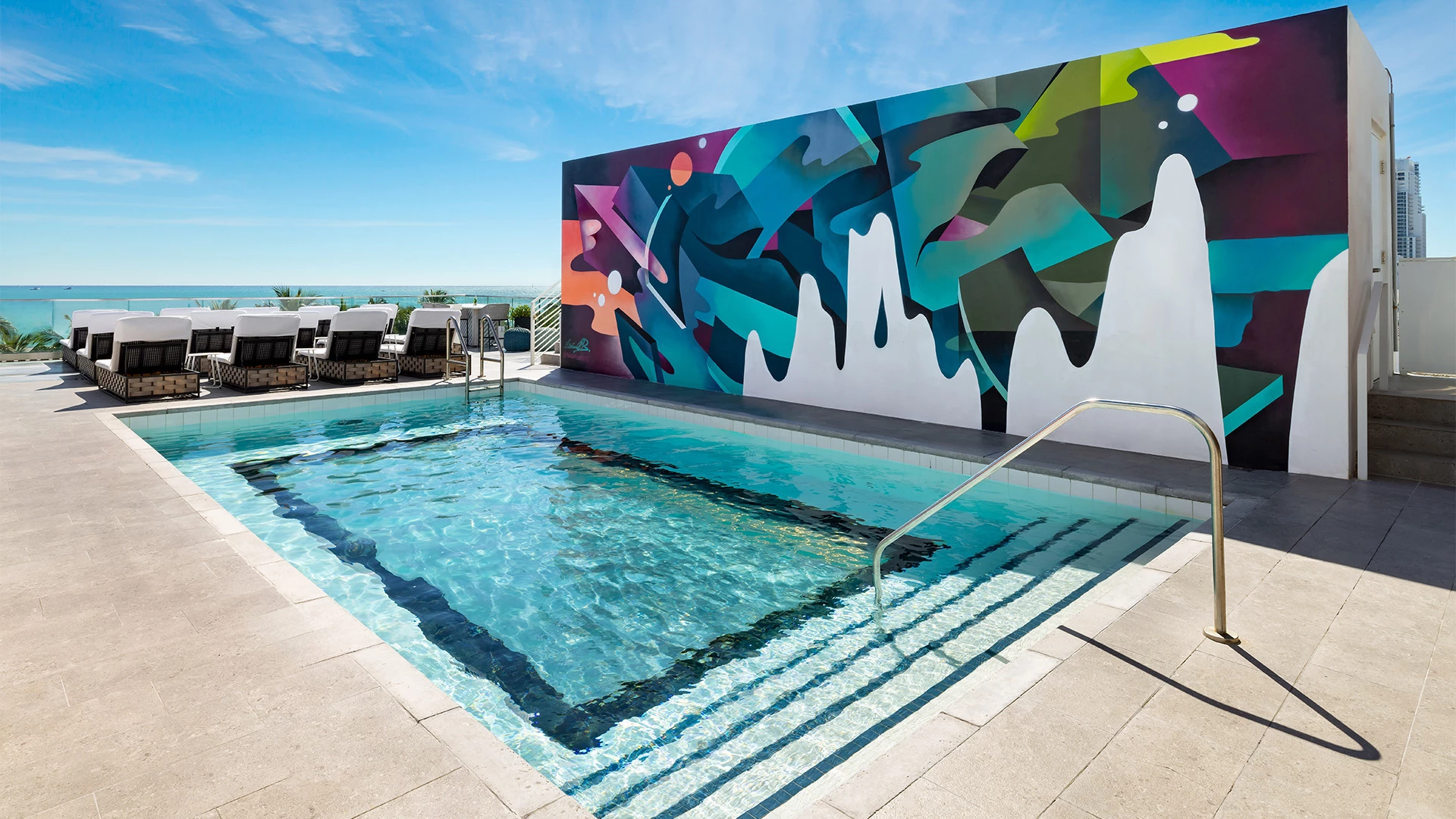 A rooftop pool with a colorful mural on one wall and lounge chairs overlooking the ocean.